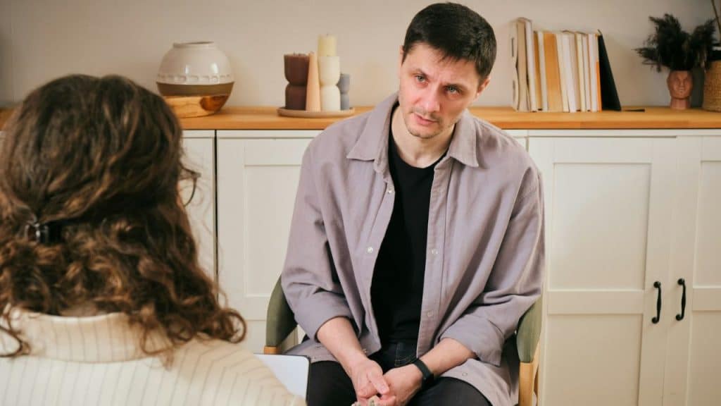 A man in a purple button-down shirt sits and speaks to a person with long hair.