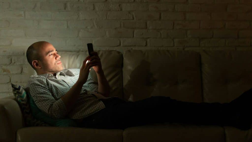 A man lies on a couch in a dark room looking at a glowing phone.