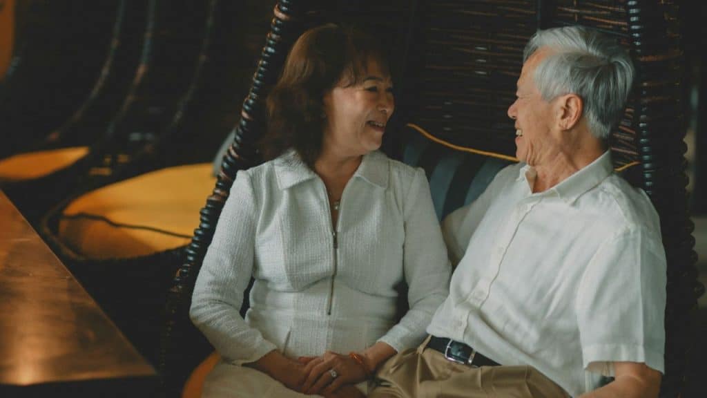An older man and woman in white shirts sit together and smile at each other.