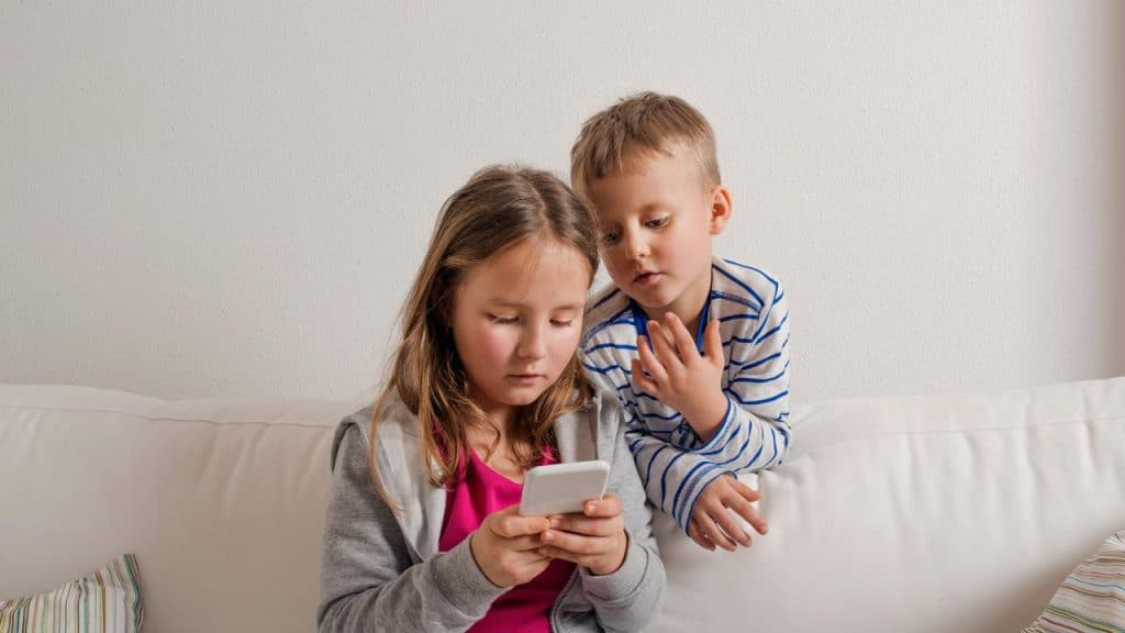 A young girl holding a smartphone while a younger boy looks over her shoulder on a couch.