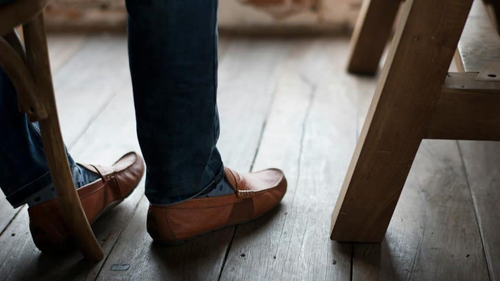 A person wearing brown loafers and jeans sitting near a wooden table.