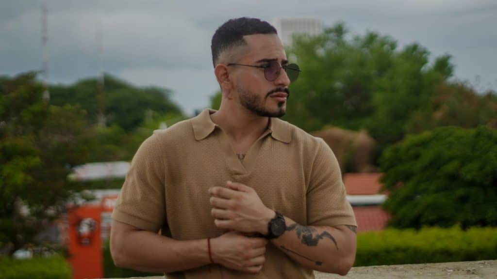 A man in a tan polo shirt and sunglasses looks toward the distance outdoors.