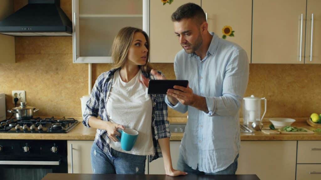 A man in a blue shirt and a woman with a mug look at a tablet in a kitchen.