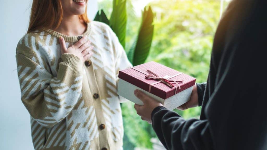 A person handing a wrapped gift box to a smiling woman indoors near a window.