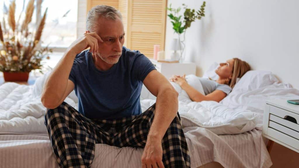 An older man sitting on a bed looking upset while a woman lies in the background.