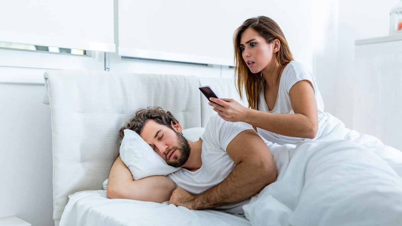 A woman sits on a bed holding a smartphone and looking shocked at a sleeping man.