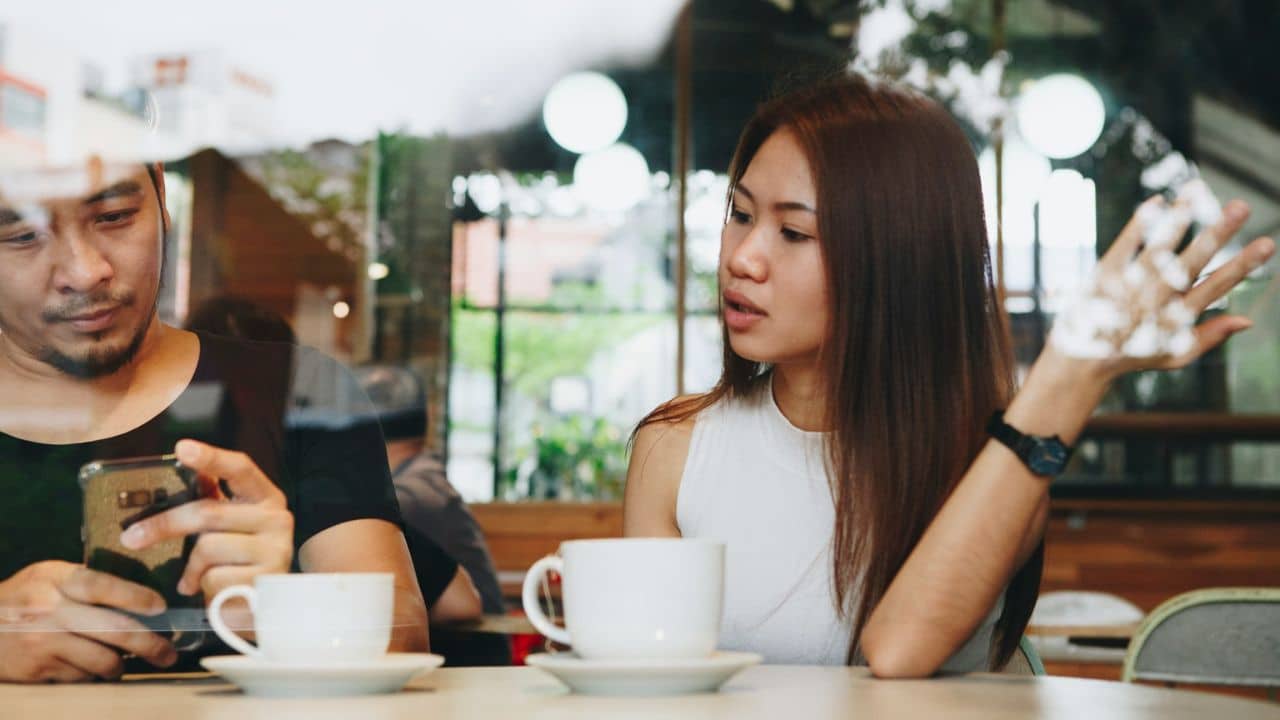 A man and woman sit at a cafe table while the man looks at a smartphone.