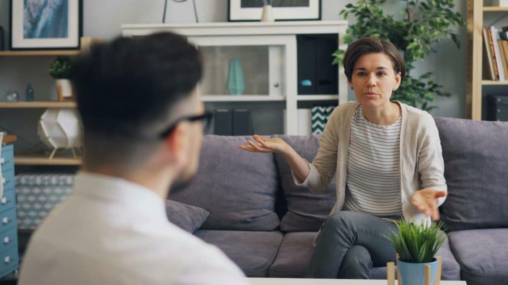 A woman sitting on a gray sofa gestures with her hands while talking to a man.