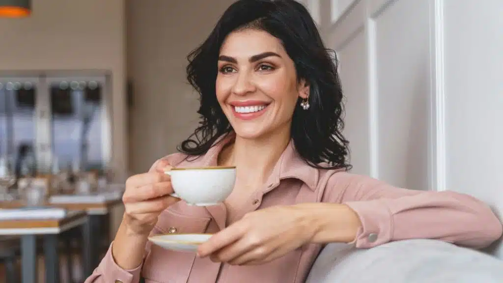 A smiling dark-haired woman in a pink shirt holds a tea cup and saucer.