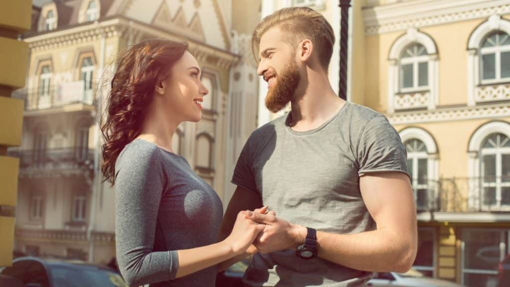A man and woman stand outside holding hands while smiling and making eye contact.
