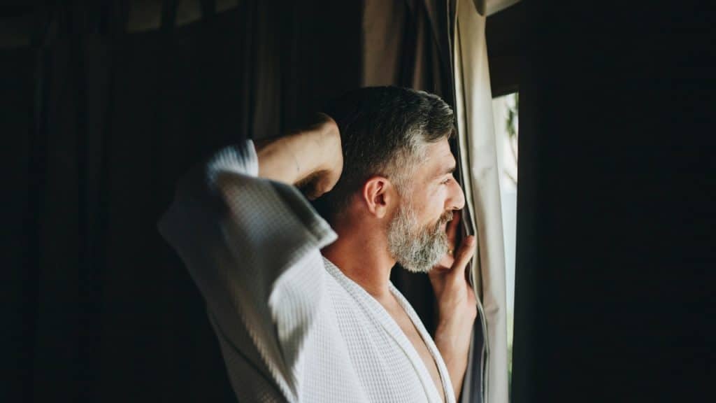 A man with a graying beard wearing a white robe peeks through dark curtains out a window.