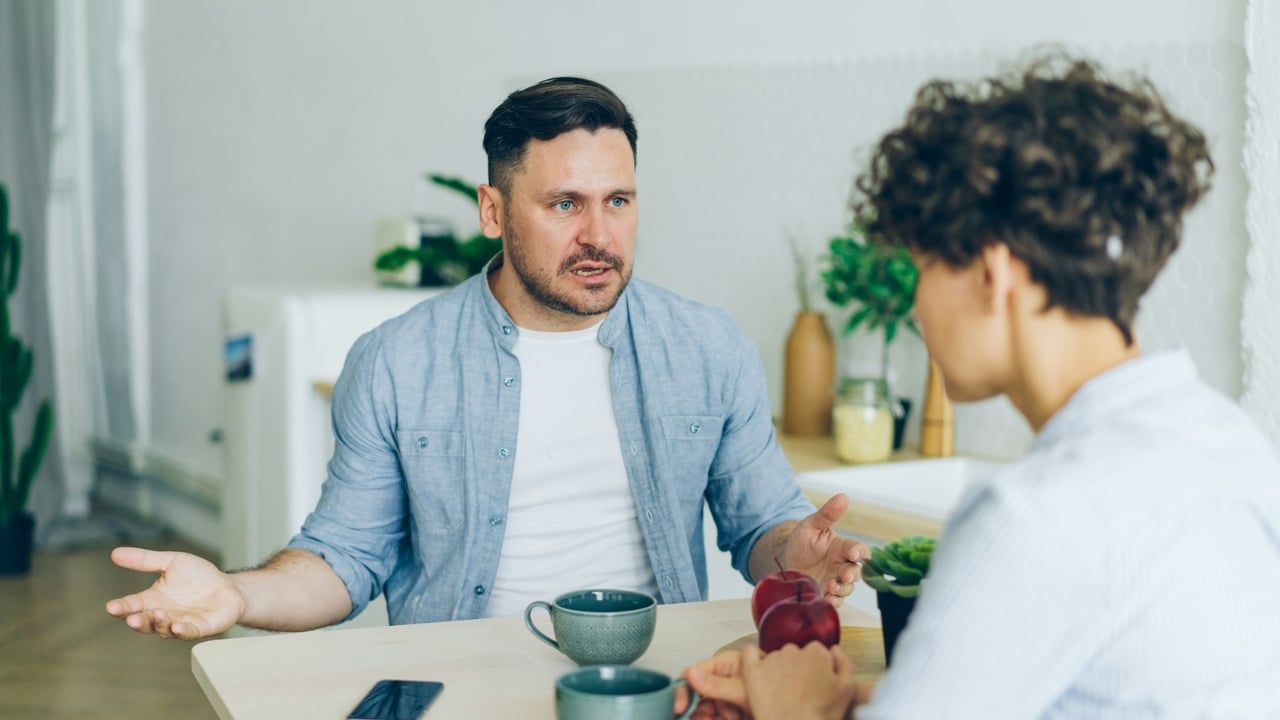 A man gestures with open palms while speaking intensely to a woman at a table.