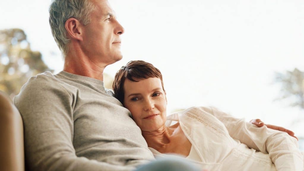A mature man and woman reclining together, with the woman resting her head on the man's chest as they both gaze thoughtfully into the distance.