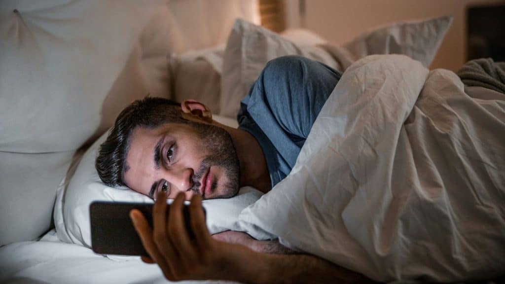 A man lies in bed in a dark room, looking at a glowing smartphone screen.