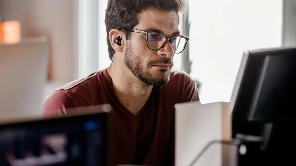 A man wearing glasses and earbuds focused on a computer screen.