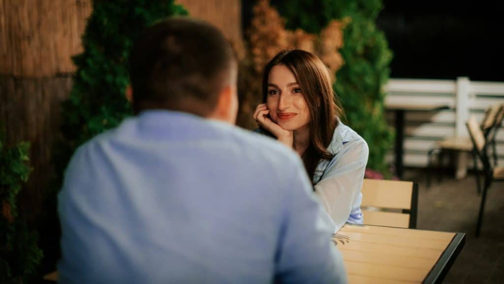 A woman rests her chin on her hand while looking at a man at an outdoor table.