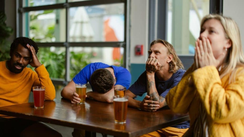 Four people sit at a table with drinks, looking tired or resting their heads.