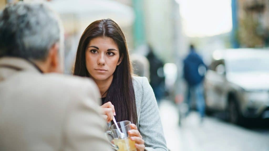 A woman with long dark hair stares blankly while sitting across from a blurred man.