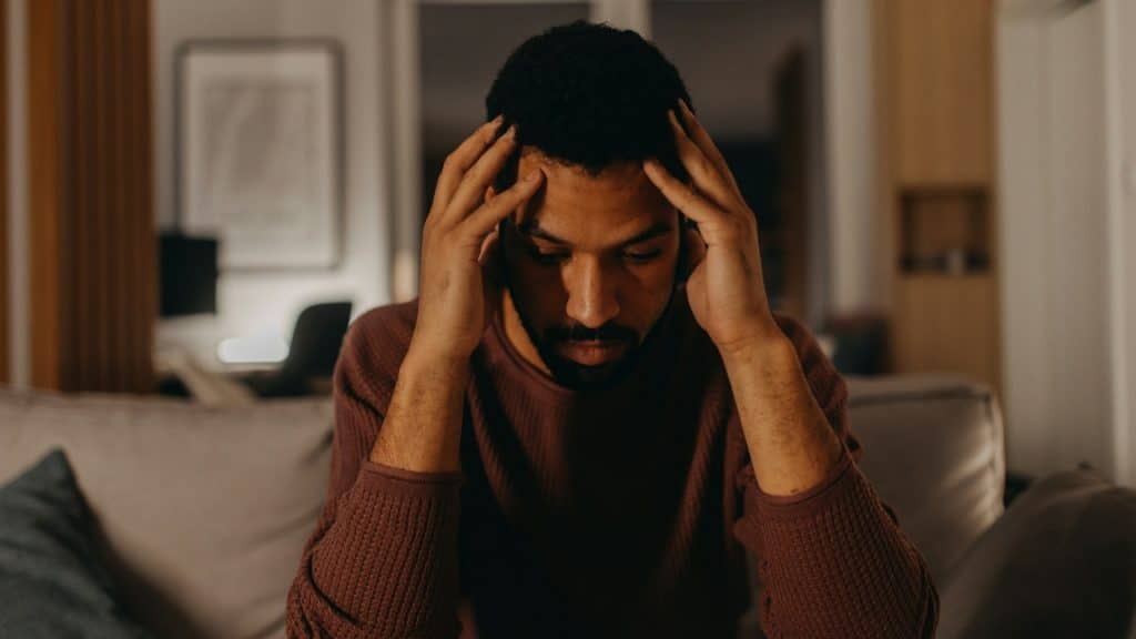 A man sits on a couch with his eyes closed, holding his head in his hands.