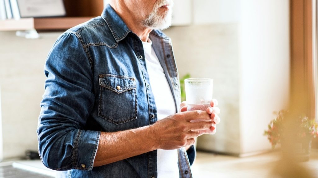 A man with a white beard wearing a denim shirt holds a glass of water.