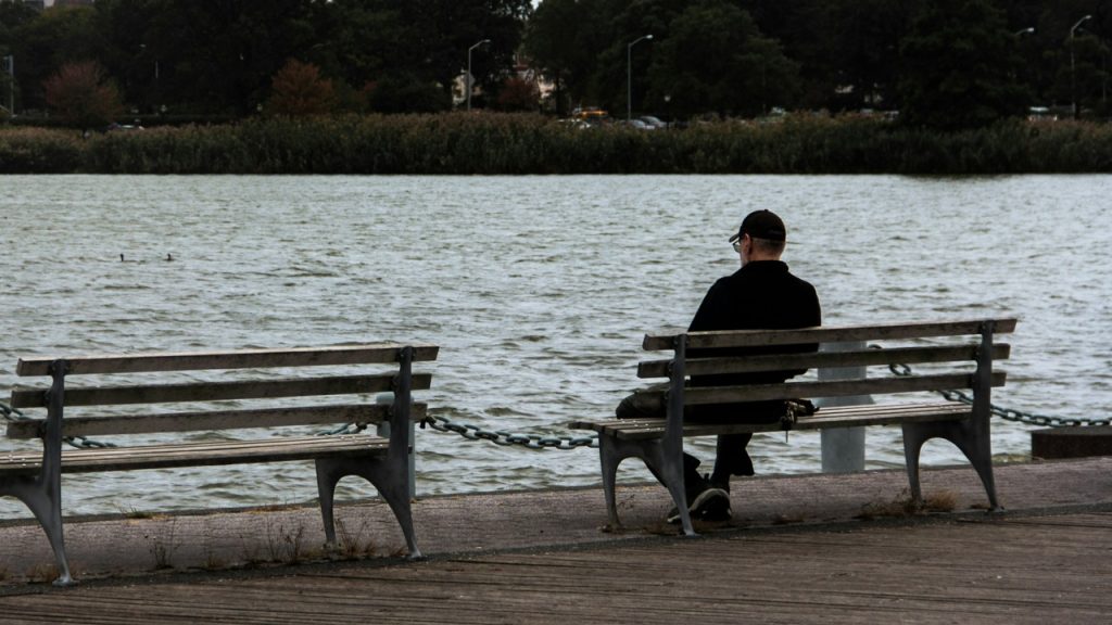 A man in dark clothing sits alone on a park bench facing a body of water.