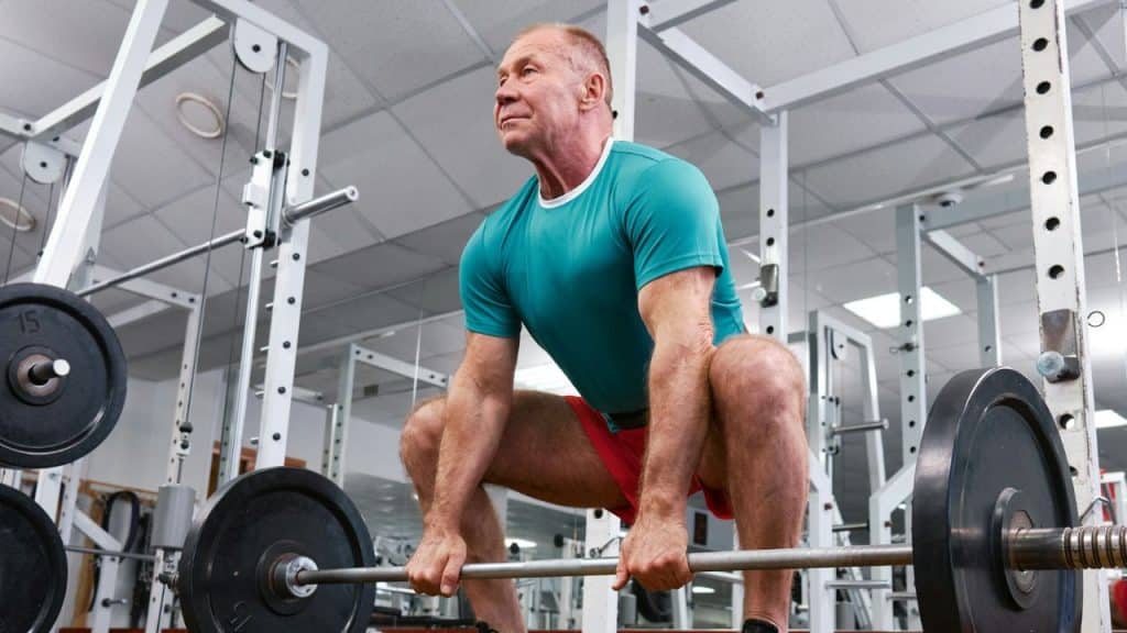 A muscular older man in a gym prepares to lift a heavy barbell from the floor.