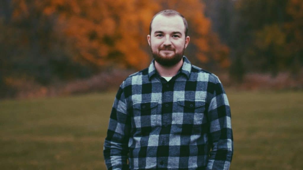 A bearded man in a plaid shirt stands in a field with autumn trees behind him.