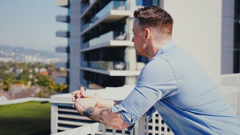 A man with tattoos on his arm leans on a balcony railing overlooking a city.