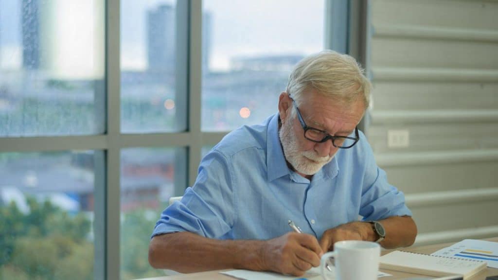 An elderly man with glasses and a beard writes at a desk next to a window.