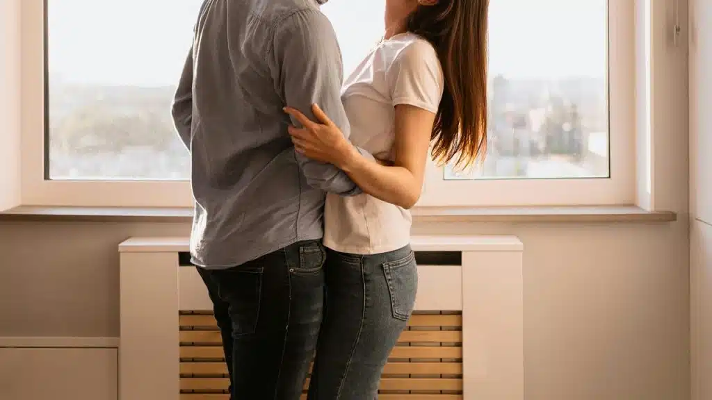 A couple embraces while standing by a sunlit window.