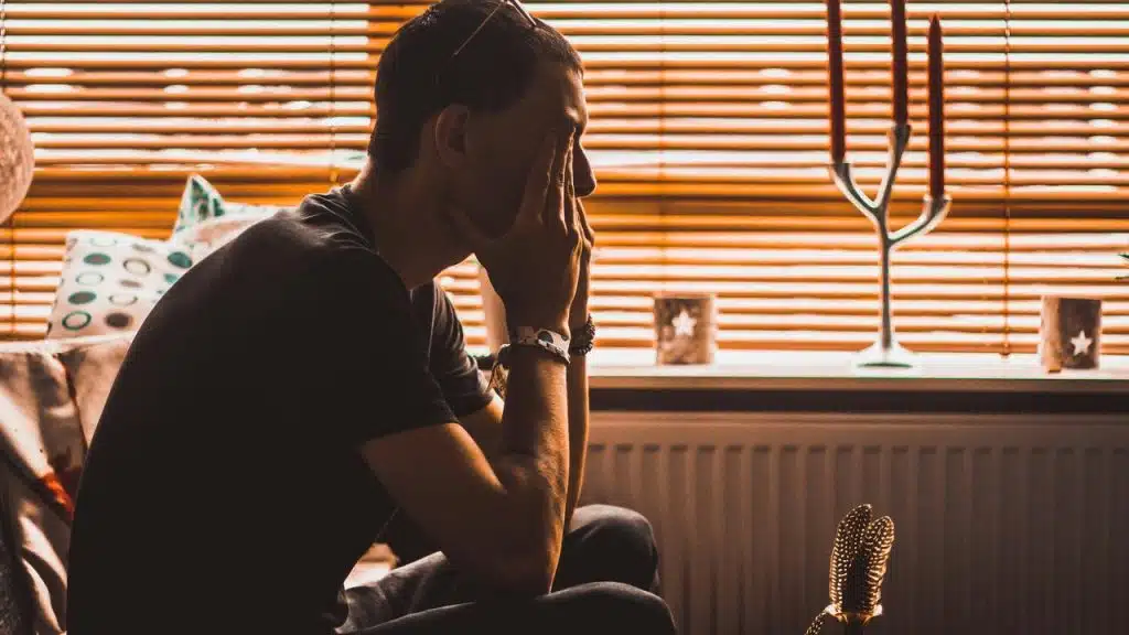 A person sitting indoors with hands on their face, appearing deep in thought by a window.
