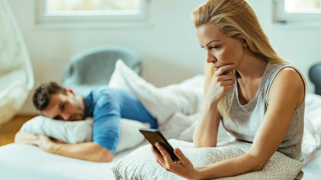 A blonde woman sits up in bed looking at a phone with a man besides.