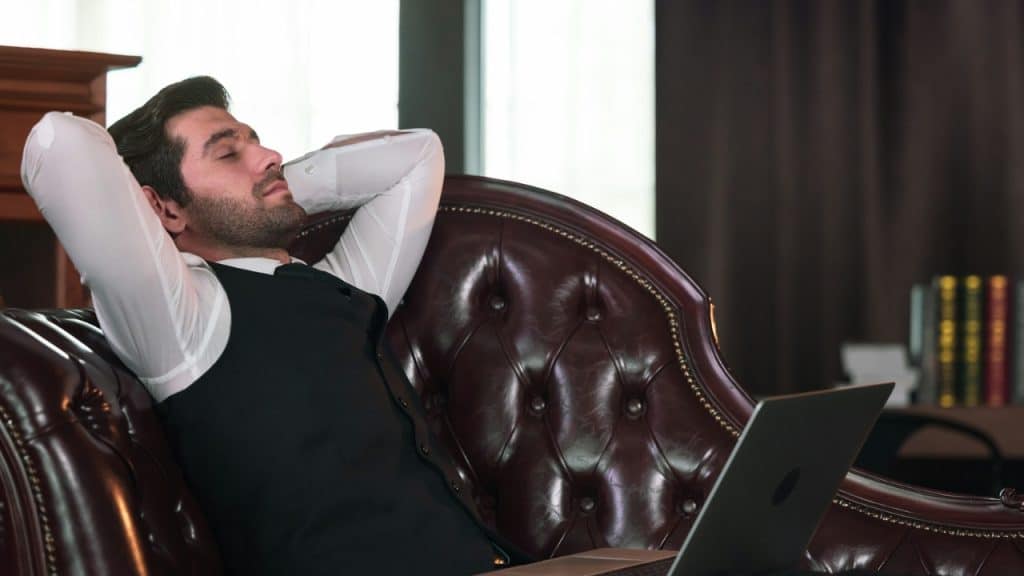 A man in a vest leans back on a leather sofa with his eyes closed.