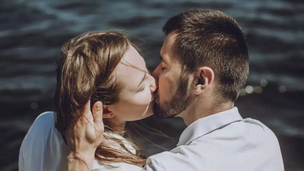 A man and woman kiss while outdoors with a body of water in the background.