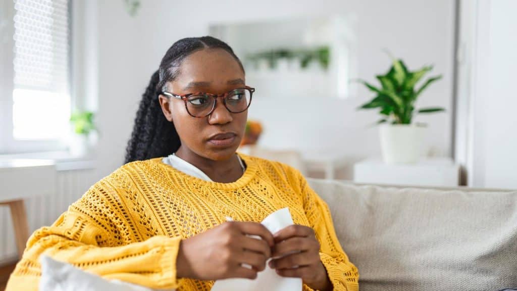 A woman sitting on a couch holding a tissue and looking concerned.