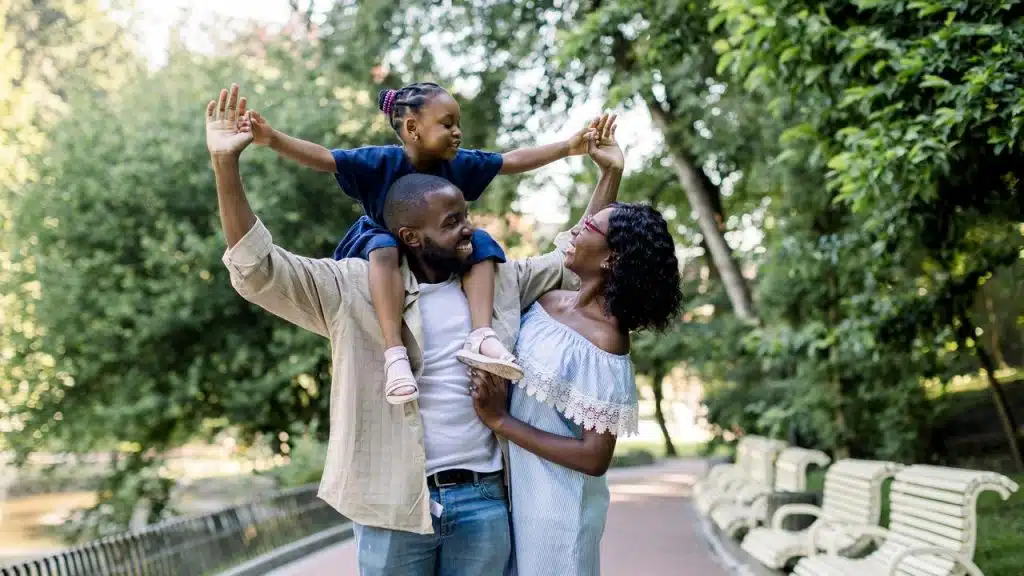 A joyful family walking in a park with a child on an adult’s shoulders.