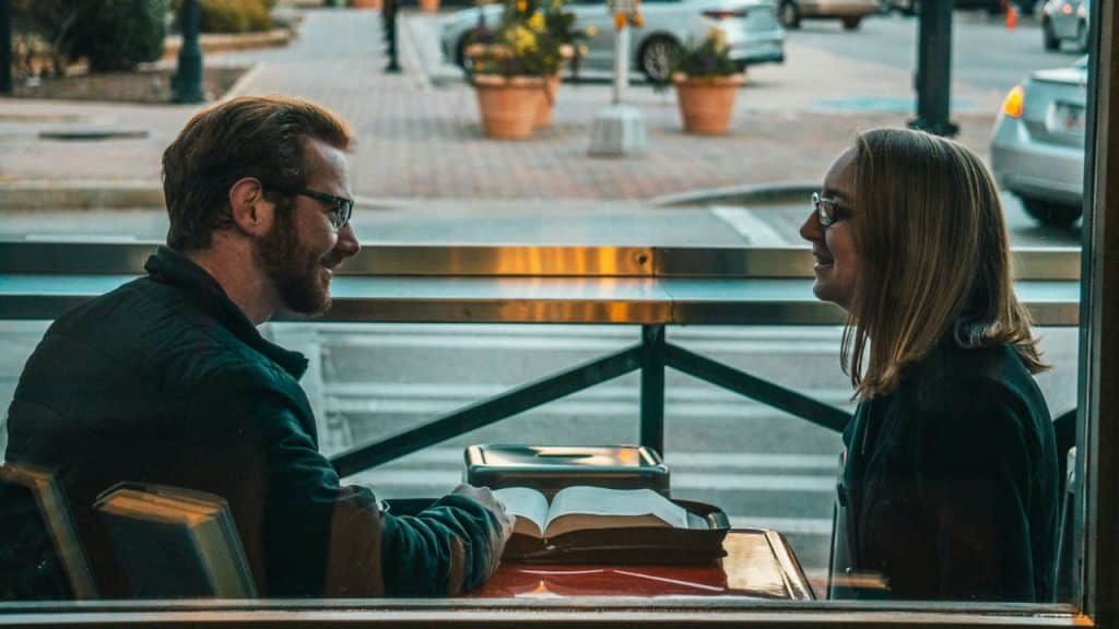 A man and a woman sit facing each other at a window counter, smiling while talking.