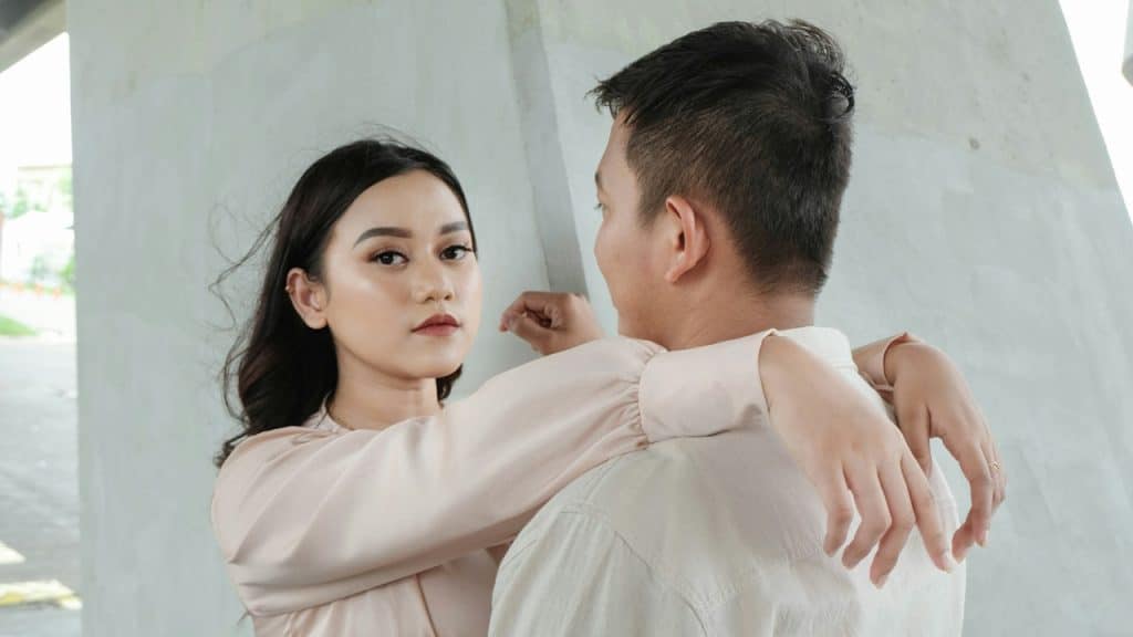 A woman looking at the camera with an inscrutable expression and resting her arms on a man’s shoulder.