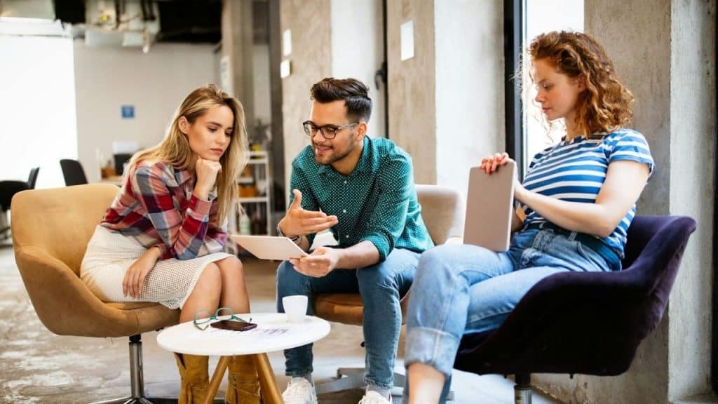 Three people sit in an office space looking at a tablet while one holds a laptop.