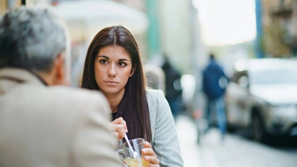 A dark-haired woman stirs a drink and looks at a person whose back is visible.