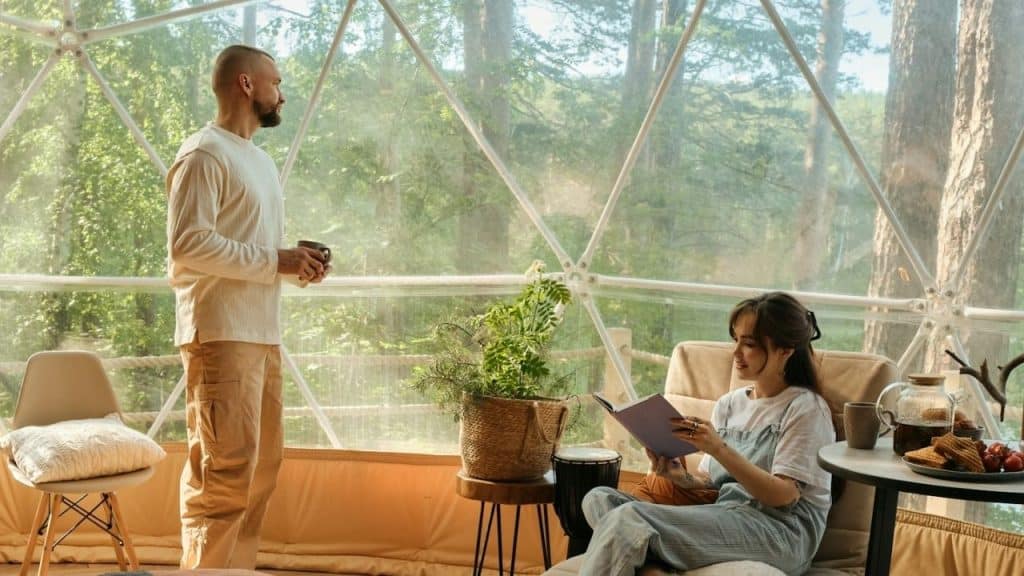 A woman reads a book while a man stands nearby looking out a window.