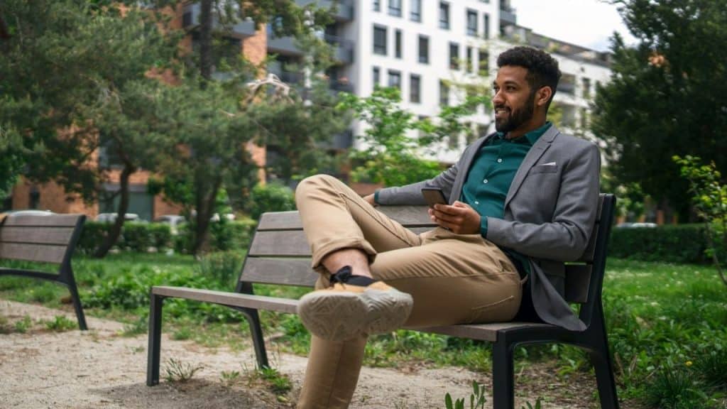 A man in a grey blazer sits on a park bench while holding a smartphone.