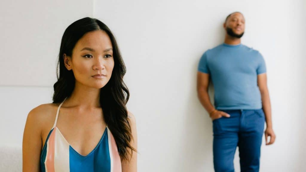 A woman looks forward while a man stands blurred in the background.