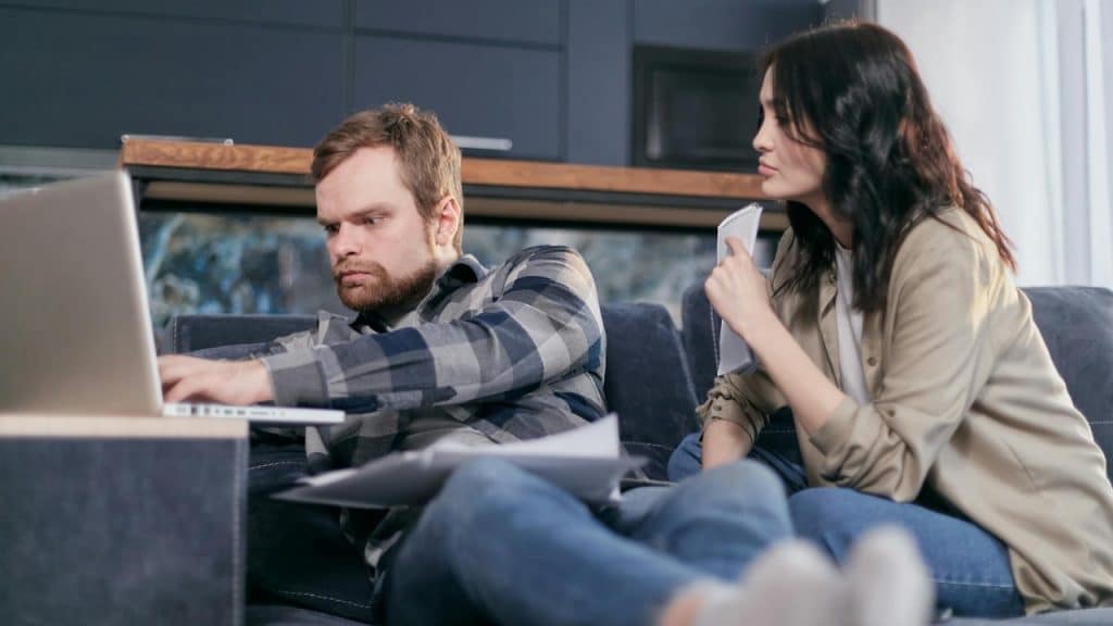 A man types on a laptop while a woman sits beside him holding a small notepad.