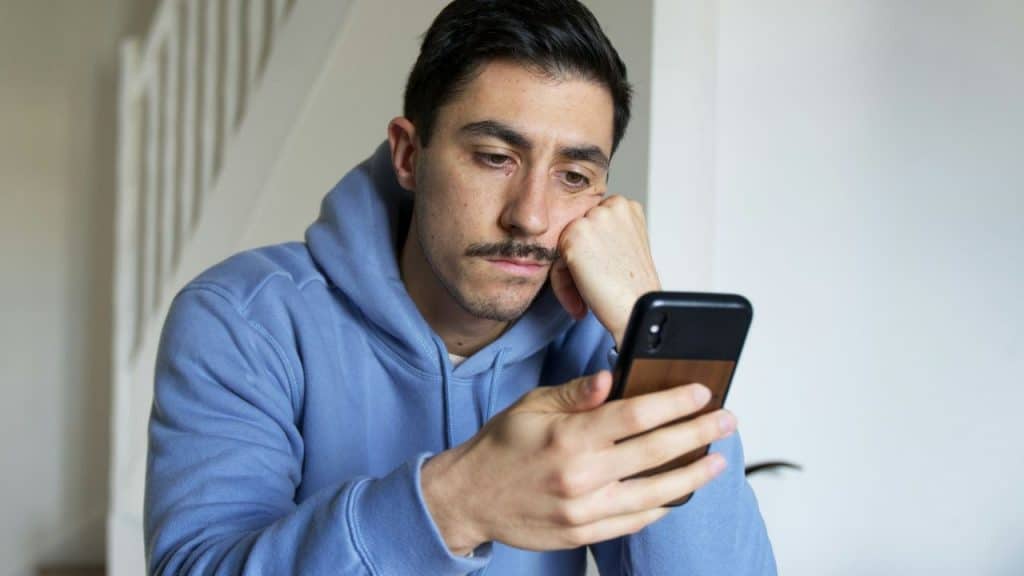 A man with a mustache in a blue hoodie rests his head on his hand while looking at a phone.