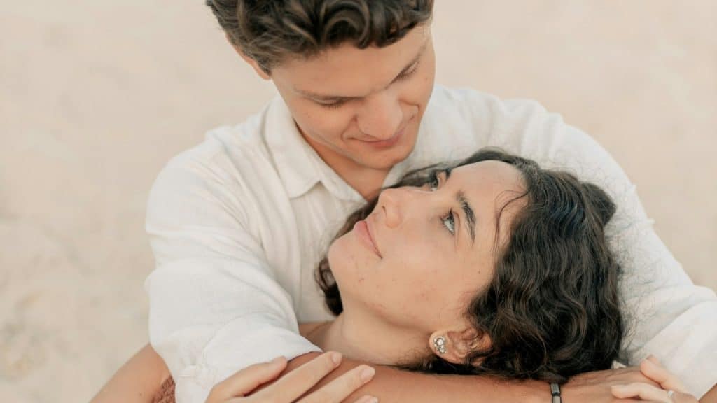 A man looks down at a woman with curly hair while embracing her from behind outdoors.