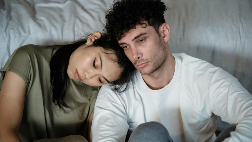 A woman rests her head on the shoulder of a man as they sit against a wall.
