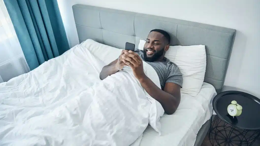 A man lies in a bed smiling while looking at a smartphone in his hands.