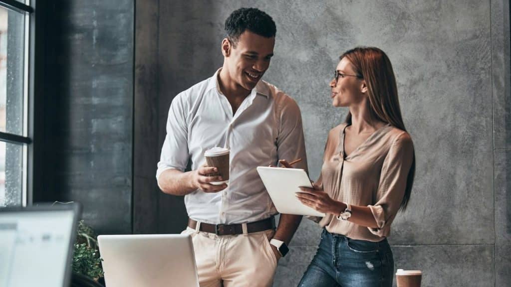 A man with a coffee cup and a woman with a tablet talk in an office.