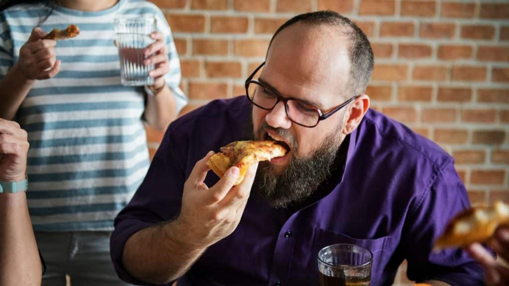 A man with a beard and glasses eats a slice of pizza at a table.